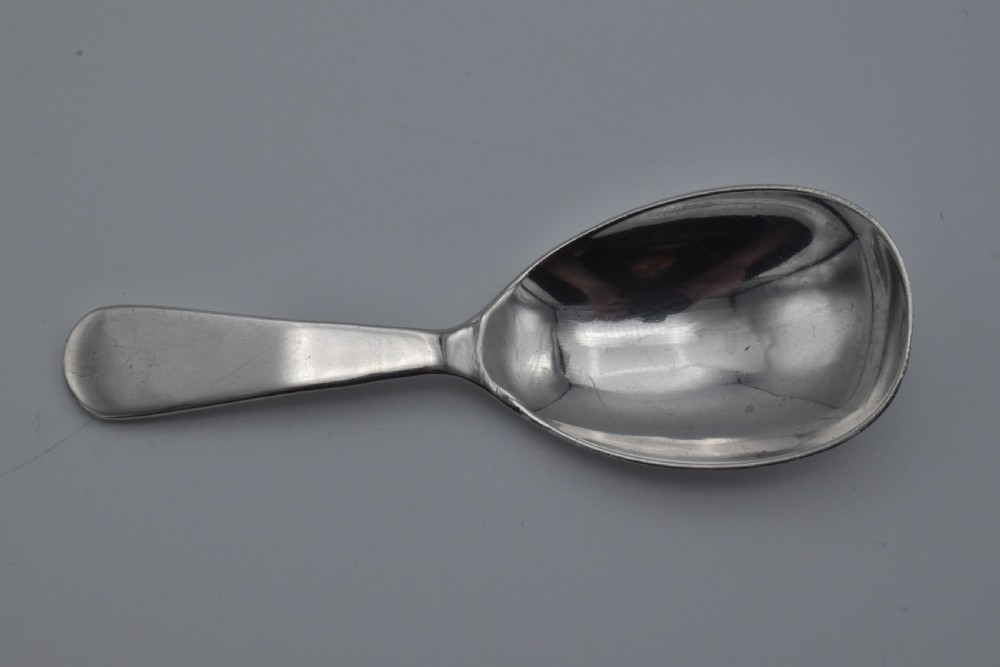 A plain sterling silver caddy spoon with a wide oval bowl and a smooth, unadorned handle resting on a neutral grey background. View IV