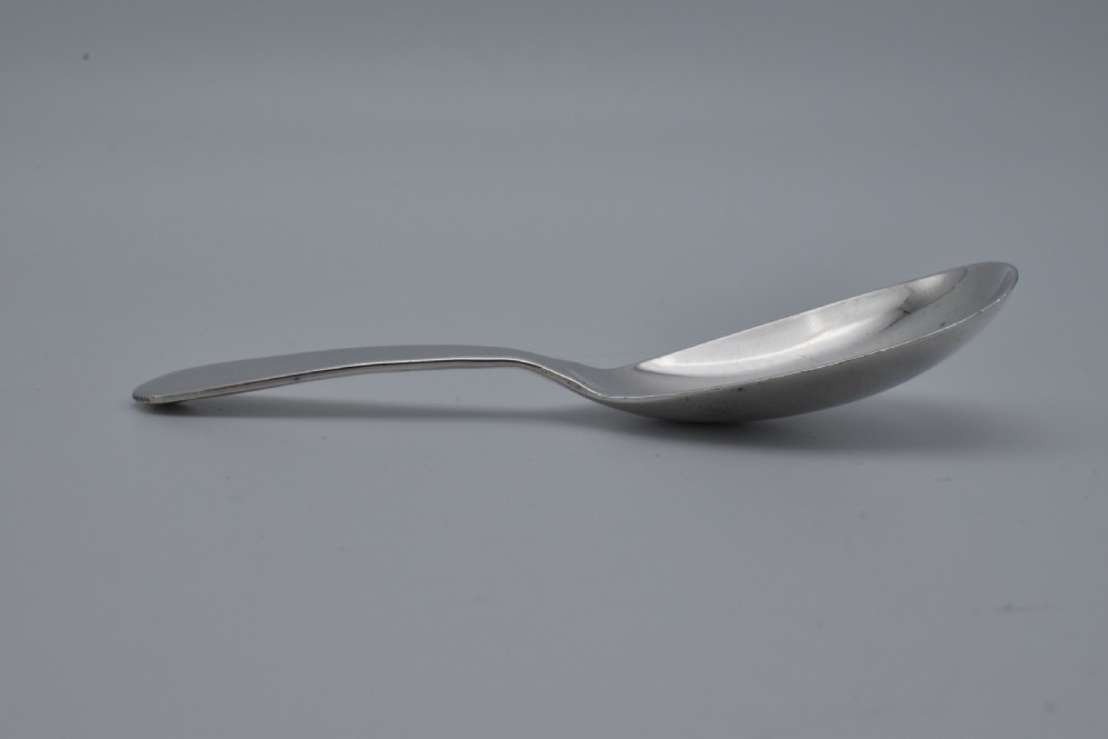 A plain sterling silver caddy spoon with a wide oval bowl and a smooth, unadorned handle resting on a neutral grey background. View V