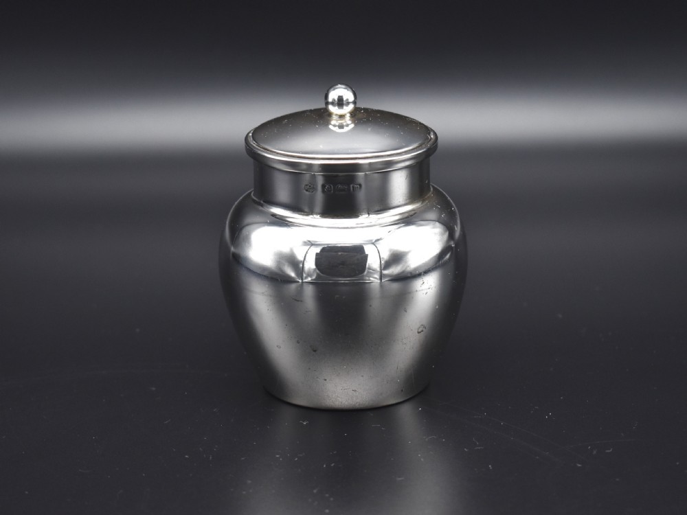 A small, bulbous sterling silver miniature tea caddy with a fitted lid, round ball finial, and visible British hallmarks stamped around the neck.