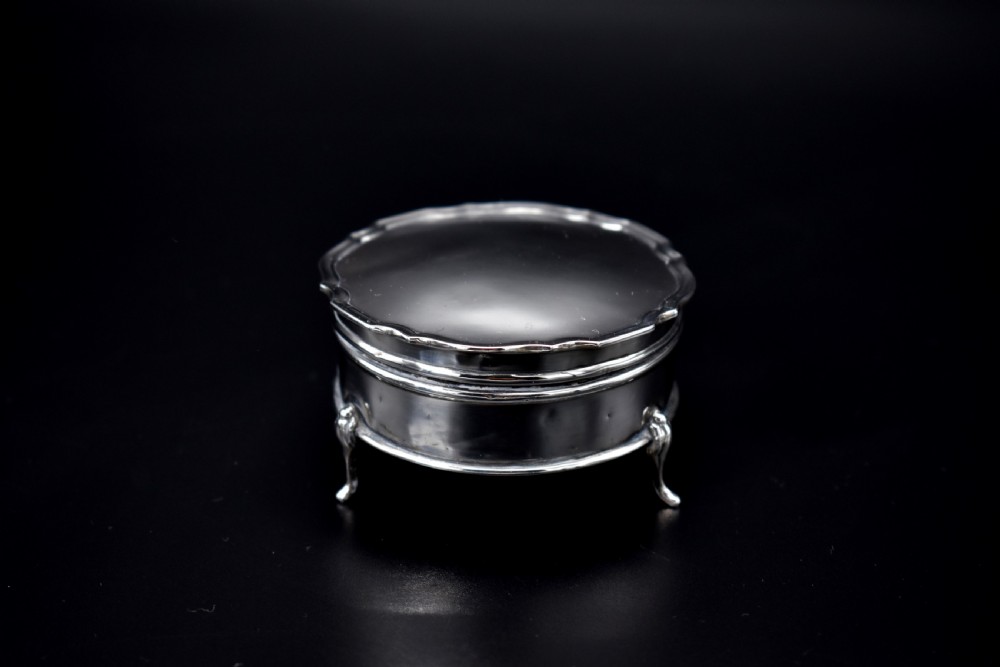 A circular sterling silver dressing table box with a shaped rim and cabriole feet displayed on a dark surface.