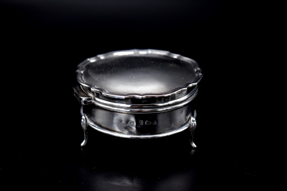 A circular sterling silver dressing table box with a shaped rim and cabriole feet displayed on a dark surface. View II