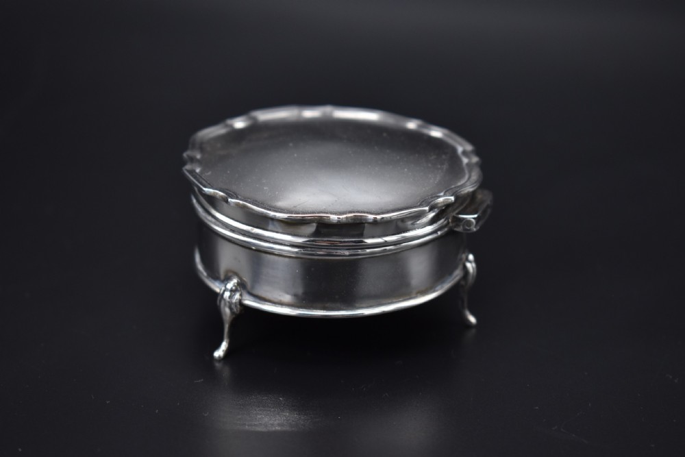 A circular sterling silver dressing table box with a shaped rim and cabriole feet displayed on a dark surface. View III