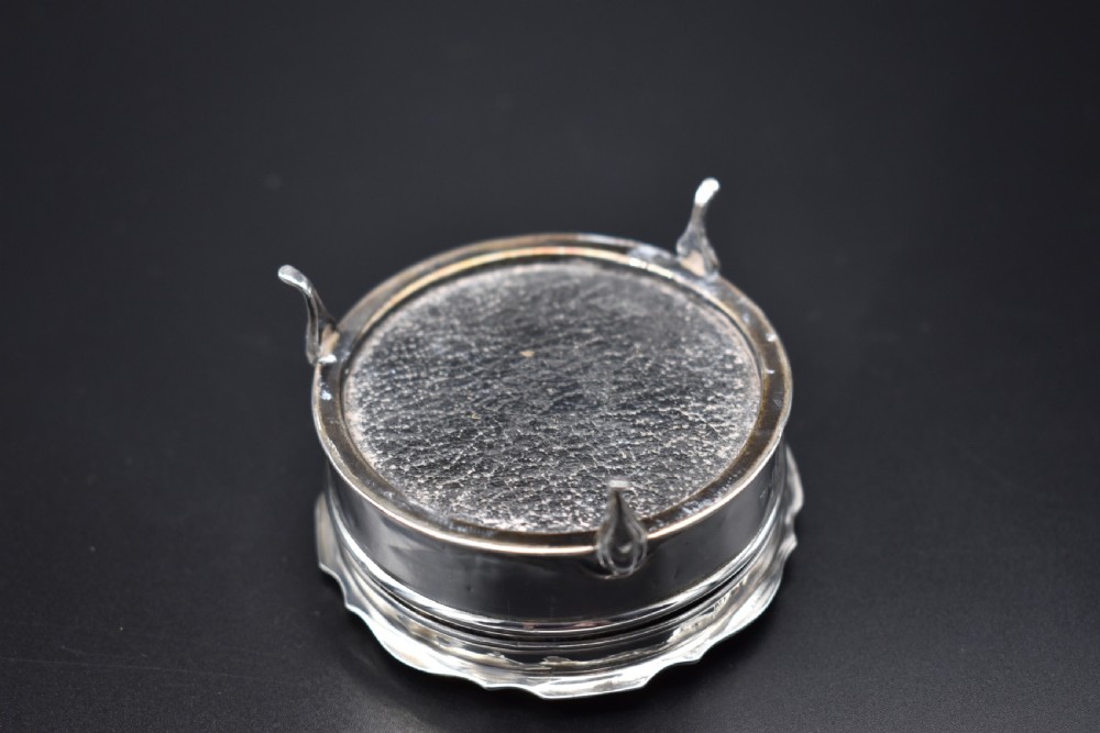 A circular sterling silver dressing table box with a shaped rim and cabriole feet displayed on a dark surface. View V