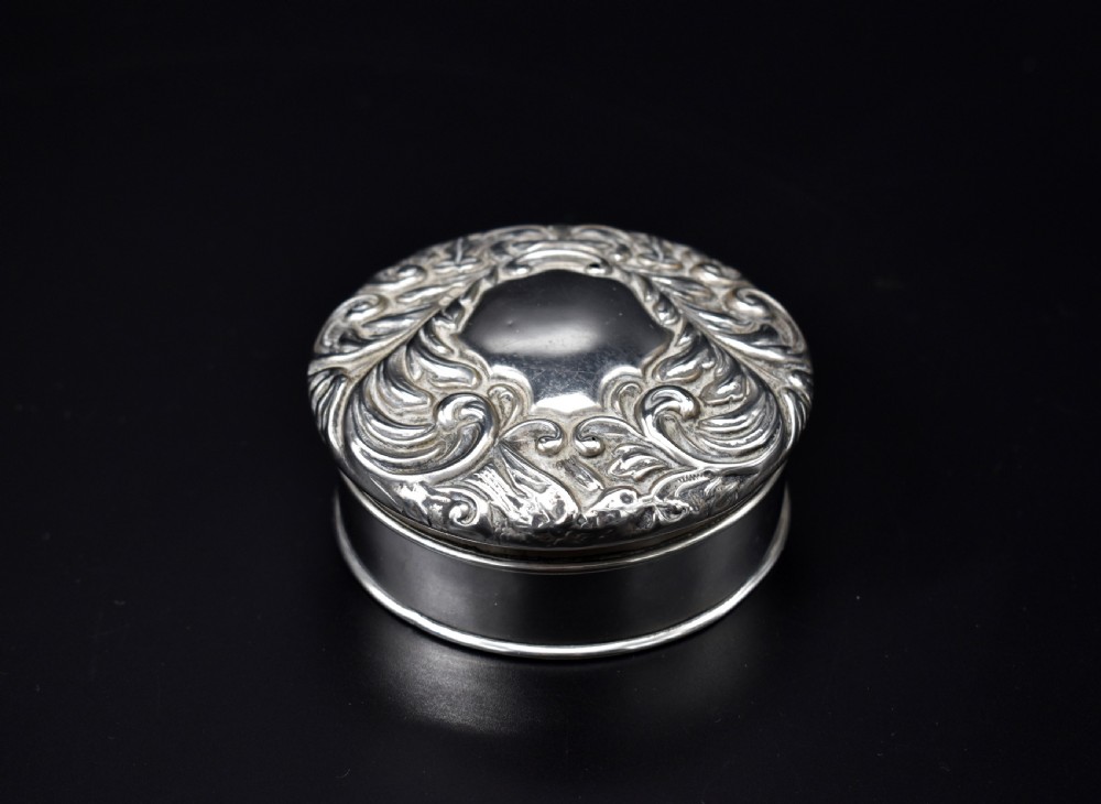 A circular Victorian sterling silver powder box featuring raised repousse scrollwork and foliage on the lid with a smooth cylindrical base.