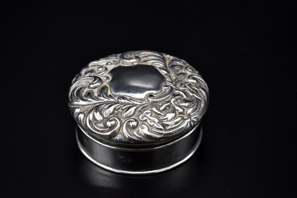 A circular Victorian sterling silver powder box featuring raised repousse scrollwork and foliage on the lid with a smooth cylindrical base. View II