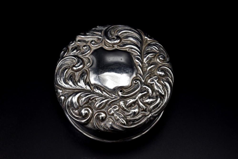 A circular Victorian sterling silver powder box featuring raised repousse scrollwork and foliage on the lid with a smooth cylindrical base. View III