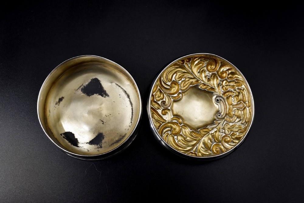 A circular Victorian sterling silver powder box featuring raised repousse scrollwork and foliage on the lid with a smooth cylindrical base. View VI