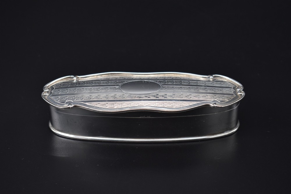 An elongated, oval Edwardian sterling silver powder box featuring engine-turned engraving on the lid and an unengraved central cartouche.