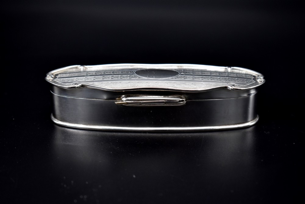 An elongated, oval Edwardian sterling silver powder box featuring engine-turned engraving on the lid and an unengraved central cartouche. View V