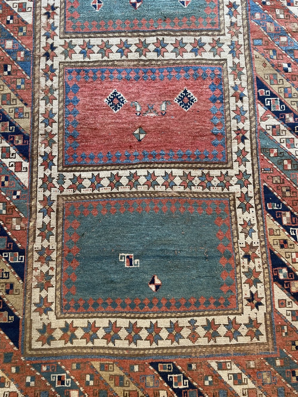 An antique hand-knotted Caucasian Bordjalou Kazak wool rug featuring three rectangular central panels surrounded by geometric diagonal borders in terracotta red, teal, cream, and blue. View IV