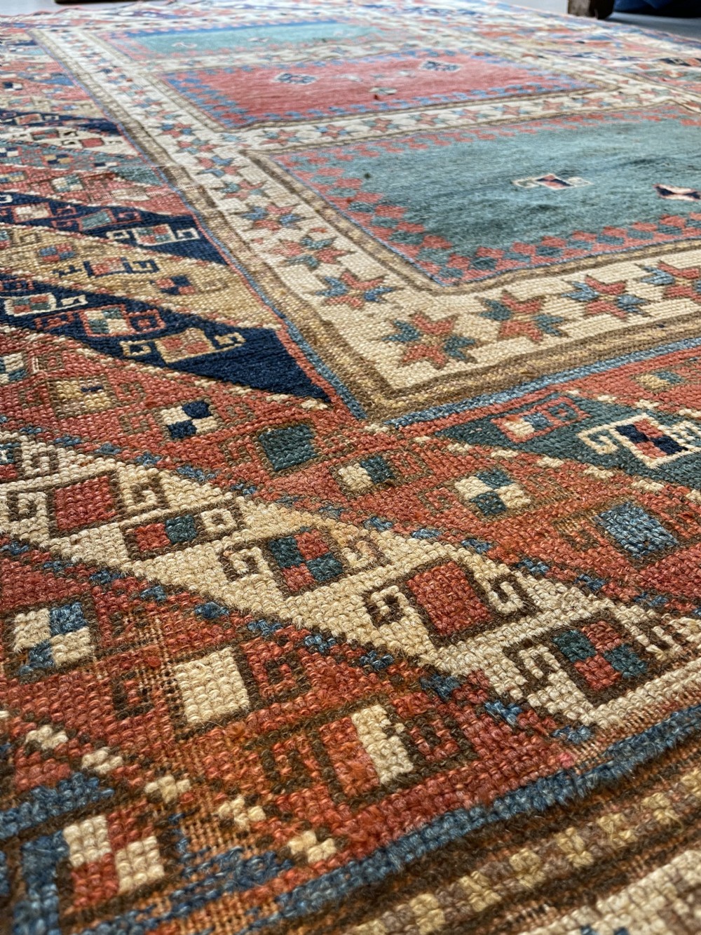 An antique hand-knotted Caucasian Bordjalou Kazak wool rug featuring three rectangular central panels surrounded by geometric diagonal borders in terracotta red, teal, cream, and blue. View V