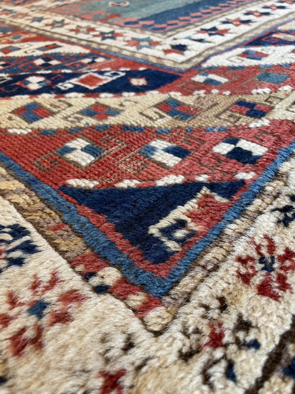 An antique hand-knotted Caucasian Bordjalou Kazak wool rug featuring three rectangular central panels surrounded by geometric diagonal borders in terracotta red, teal, cream, and blue. View VI