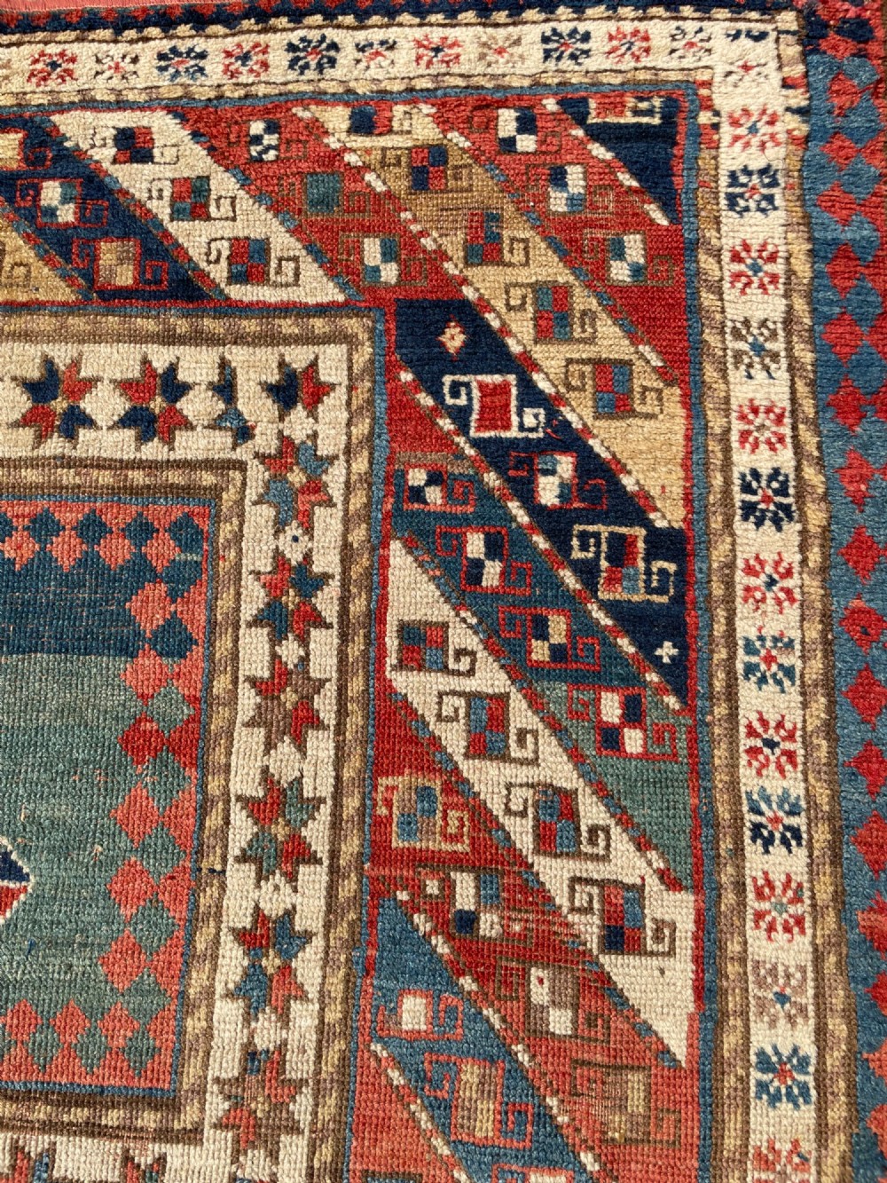 An antique hand-knotted Caucasian Bordjalou Kazak wool rug featuring three rectangular central panels surrounded by geometric diagonal borders in terracotta red, teal, cream, and blue. View VII
