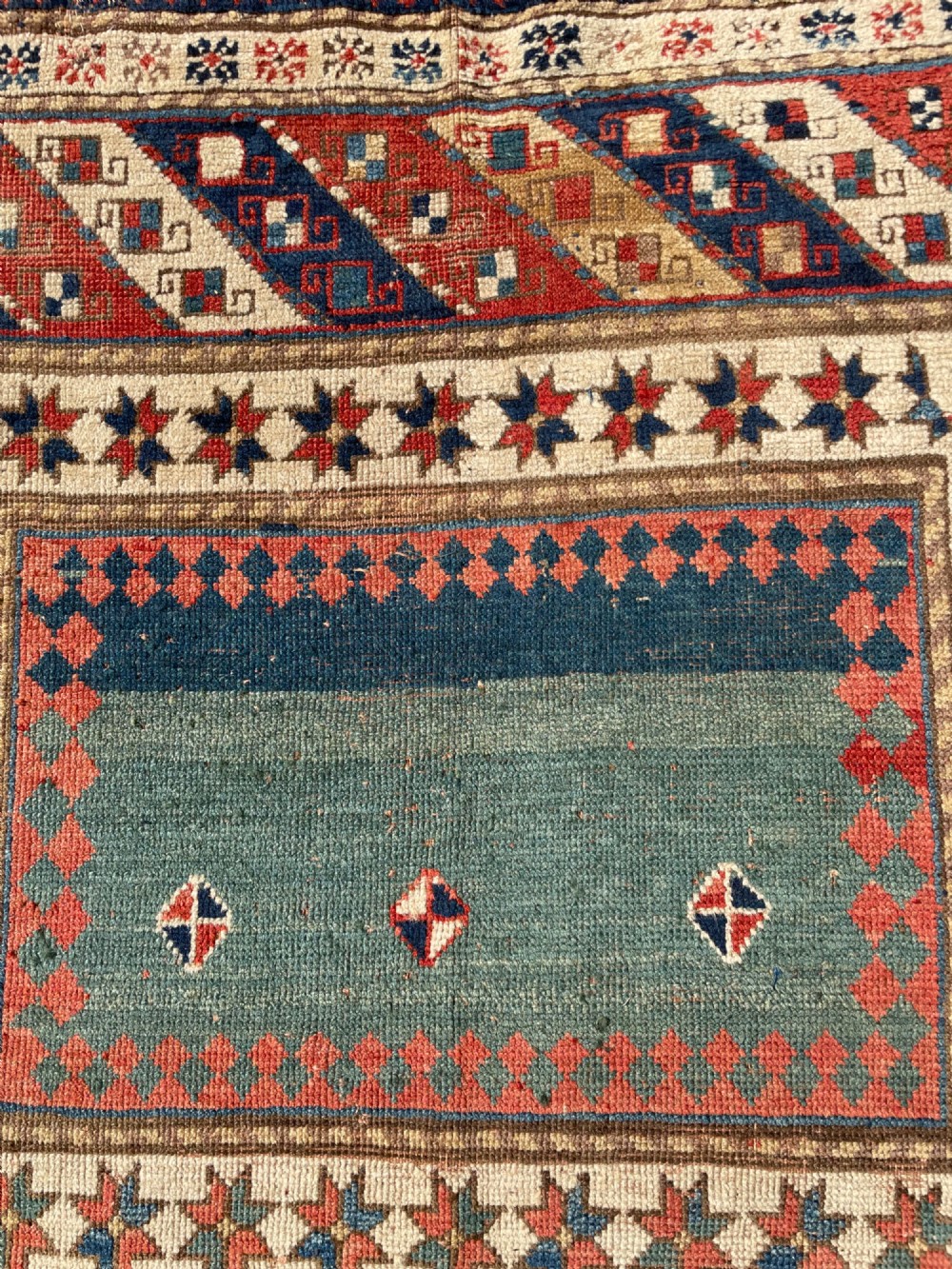 An antique hand-knotted Caucasian Bordjalou Kazak wool rug featuring three rectangular central panels surrounded by geometric diagonal borders in terracotta red, teal, cream, and blue. View VIII
