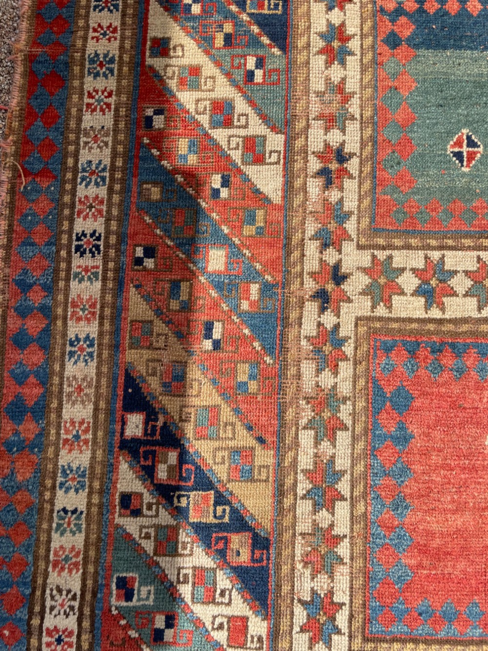An antique hand-knotted Caucasian Bordjalou Kazak wool rug featuring three rectangular central panels surrounded by geometric diagonal borders in terracotta red, teal, cream, and blue. View IX