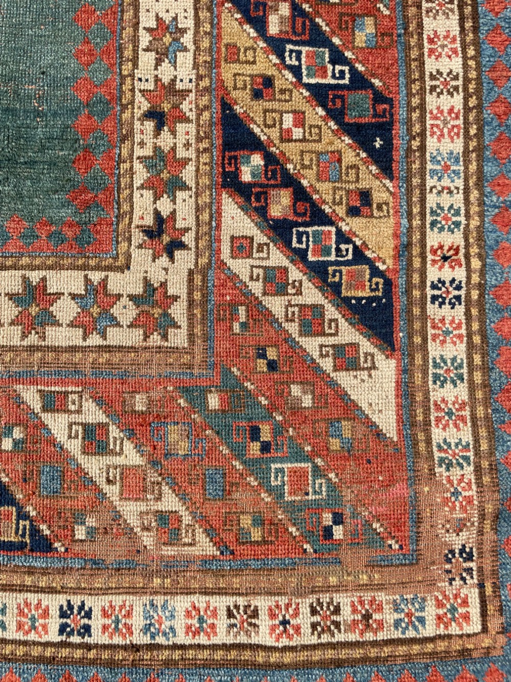 An antique hand-knotted Caucasian Bordjalou Kazak wool rug featuring three rectangular central panels surrounded by geometric diagonal borders in terracotta red, teal, cream, and blue. View XII