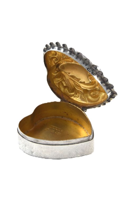 fig 1. A heart-shaped Victorian sterling silver pill box featuring elaborate repousse scrollwork on the lid, showing an open interior with gold gilding.