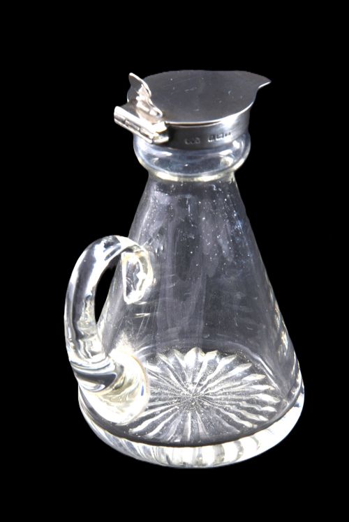 fig 1. A conical clear glass whisky noggin featuring a starburst cut base, a curved glass handle, and a silver collar with a hinged lid.