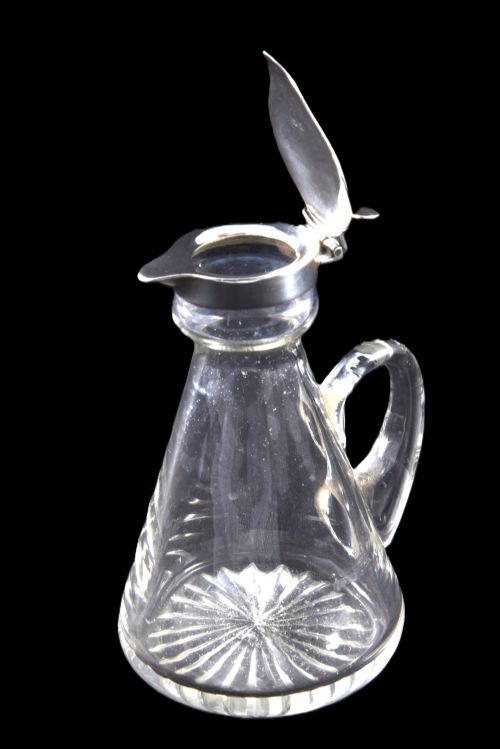 fig 2. A conical clear glass whisky noggin featuring a starburst cut base, a curved glass handle, and a silver collar with a hinged lid.