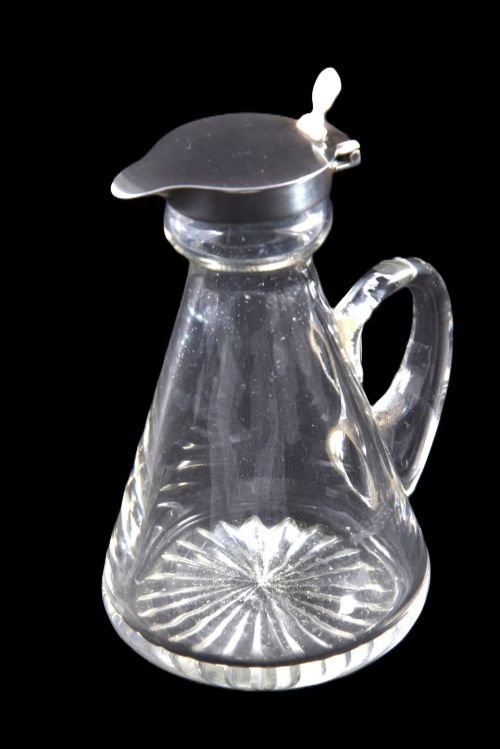 fig 3. A conical clear glass whisky noggin featuring a starburst cut base, a curved glass handle, and a silver collar with a hinged lid.