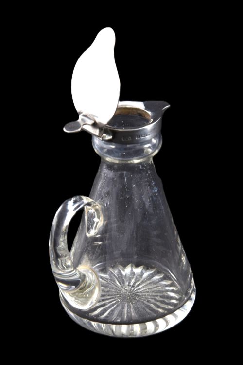fig 4. A conical clear glass whisky noggin featuring a starburst cut base, a curved glass handle, and a silver collar with a hinged lid.