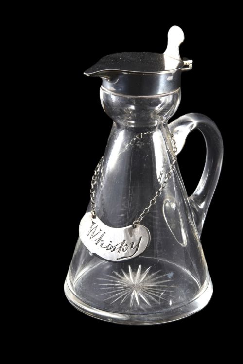 fig 1. A conical clear glass whisky noggin with a starburst cut base, fitted with a silver hinged lid and an engraved silver Whisky label hanging from a chain around its neck.