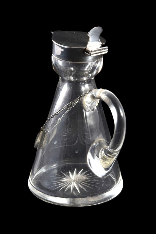 fig 2. A conical clear glass whisky noggin with a starburst cut base, fitted with a silver hinged lid and an engraved silver Whisky label hanging from a chain around its neck.