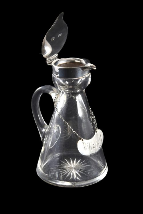 fig 4. A conical clear glass whisky noggin with a starburst cut base, fitted with a silver hinged lid and an engraved silver Whisky label hanging from a chain around its neck.
