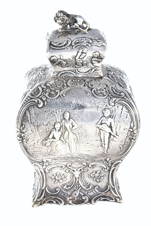 fig 1. A Victorian sterling silver tea caddy featuring a shaped bombe body with detailed repousse pastoral scenes and a lion finial on the lid.