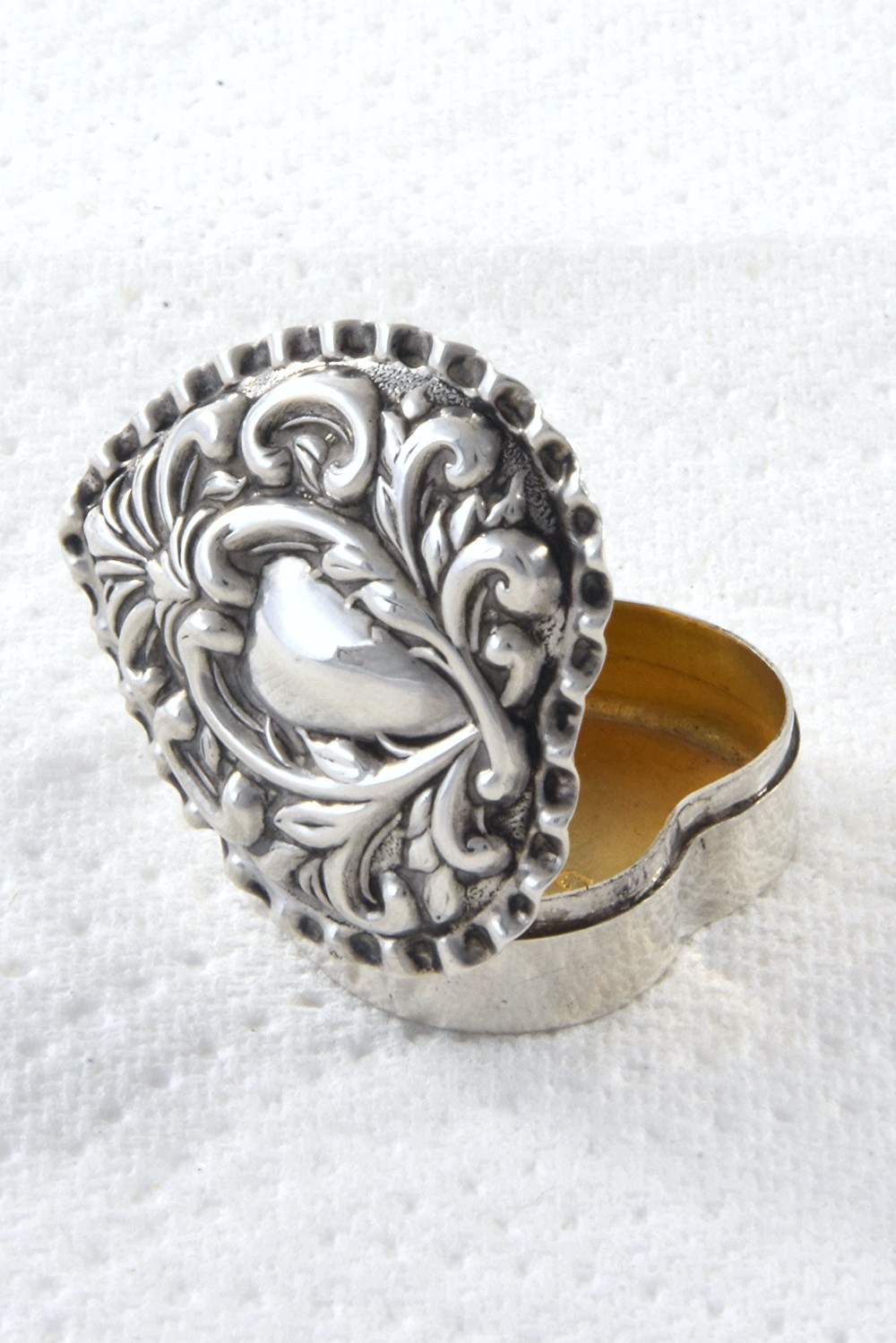 A heart-shaped Victorian sterling silver pill box featuring elaborate repousse scrollwork on the lid, showing an open interior with gold gilding.