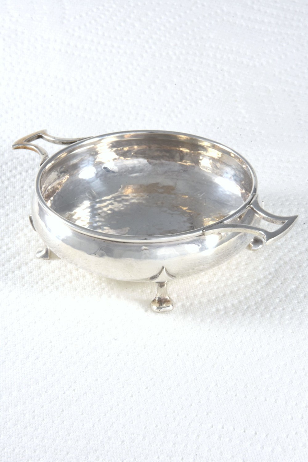 A small round George V sterling silver trinket dish featuring twin side handles, planished surface texture, and three small feet. View II