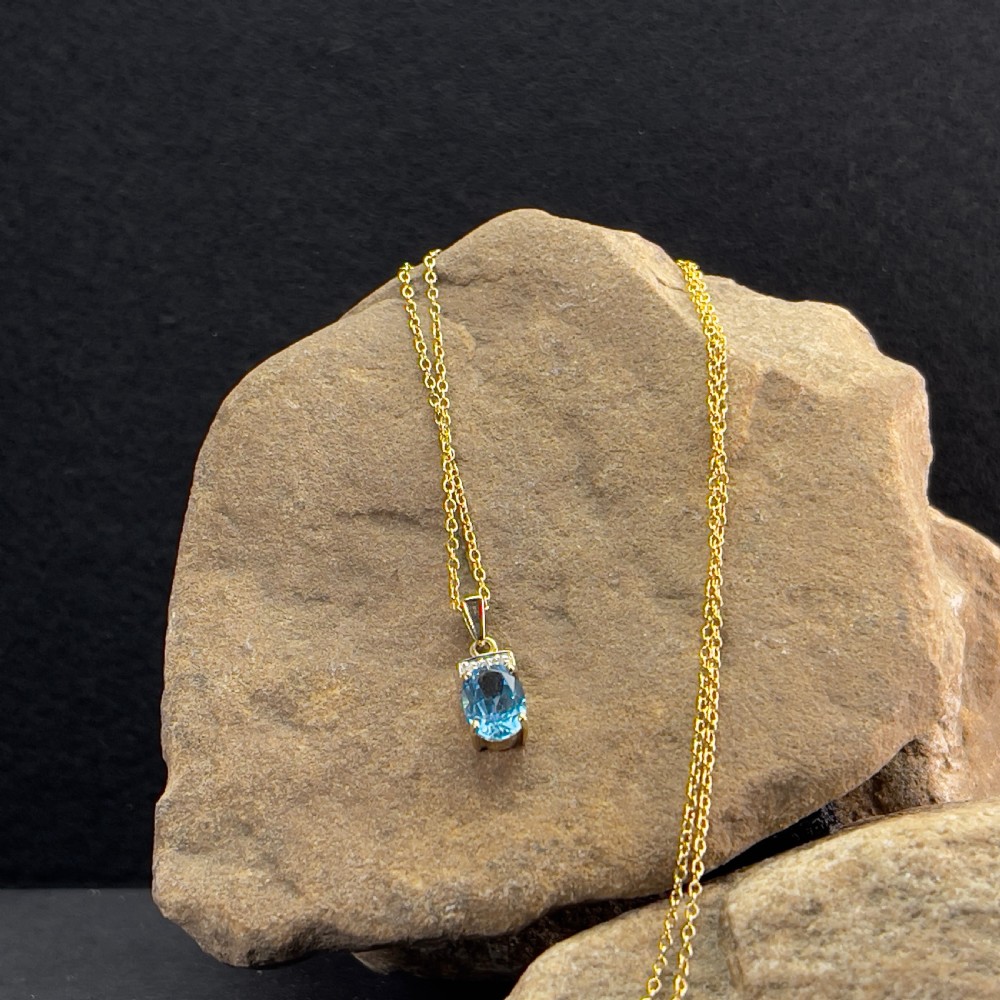 A 9ct yellow gold pendant featuring an oval faceted light blue topaz beneath a small diamond-set bar, suspended from a fine gold chain. View III
