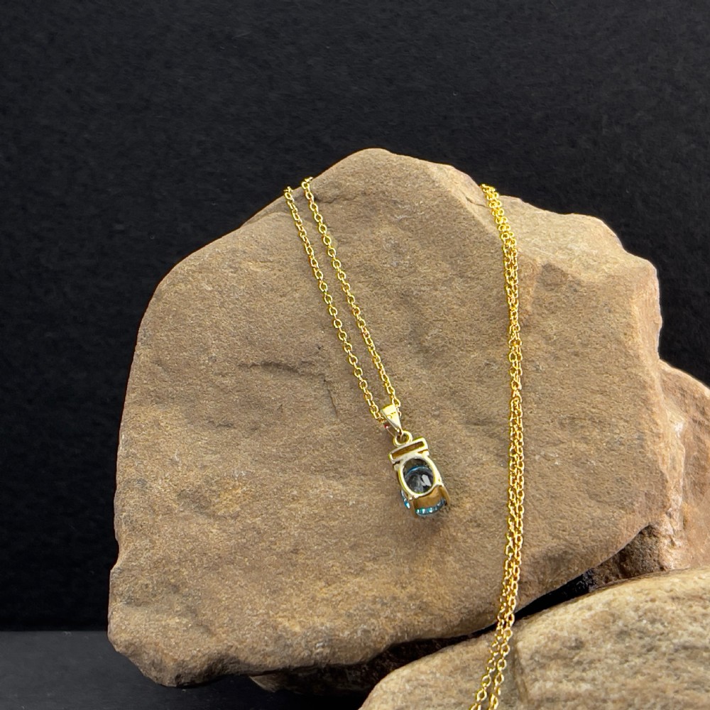 A 9ct yellow gold pendant featuring an oval faceted light blue topaz beneath a small diamond-set bar, suspended from a fine gold chain. View IV
