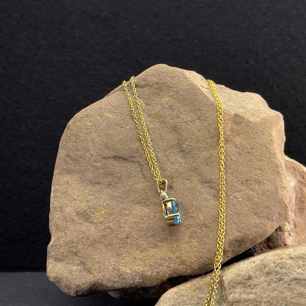 A 9ct yellow gold pendant featuring an oval faceted light blue topaz beneath a small diamond-set bar, suspended from a fine gold chain. View V