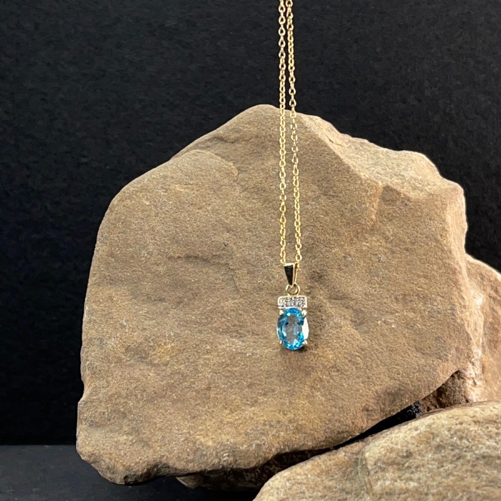 A 9ct yellow gold pendant featuring an oval faceted light blue topaz beneath a small diamond-set bar, suspended from a fine gold chain. View VI