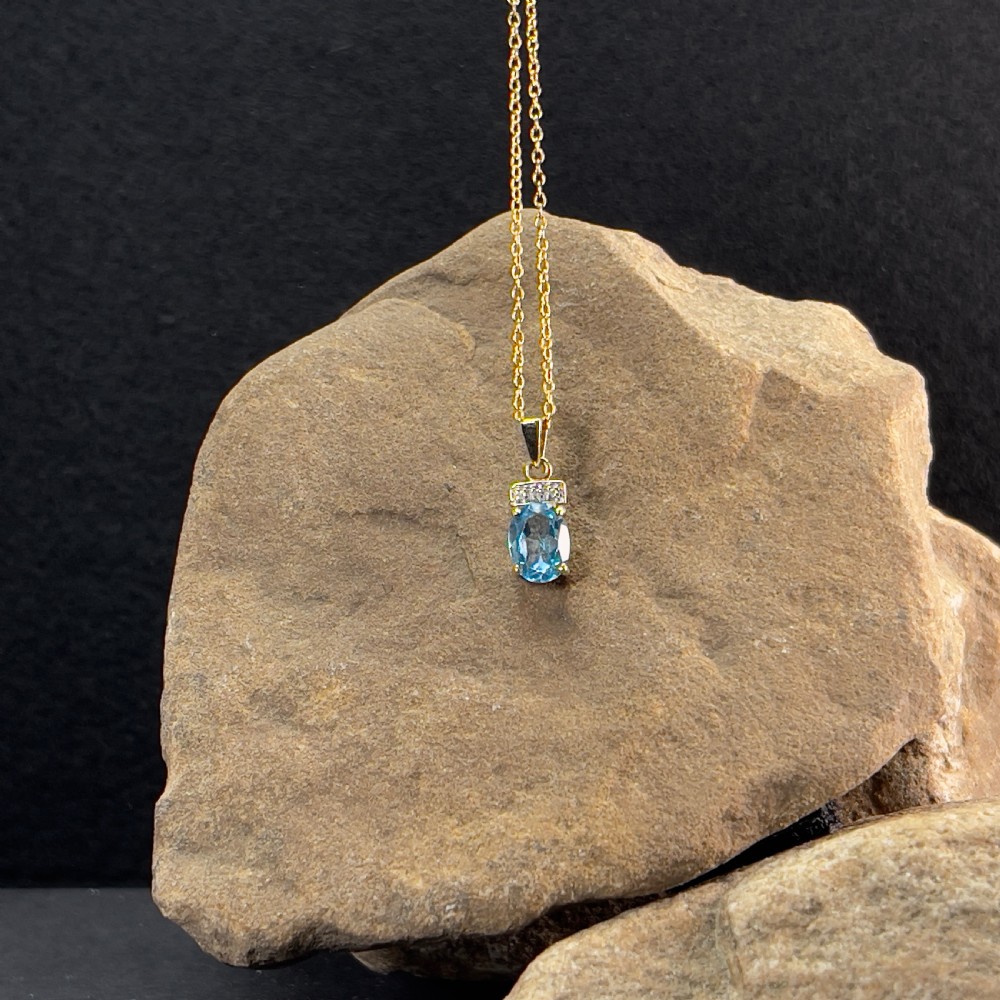 A 9ct yellow gold pendant featuring an oval faceted light blue topaz beneath a small diamond-set bar, suspended from a fine gold chain. View VII