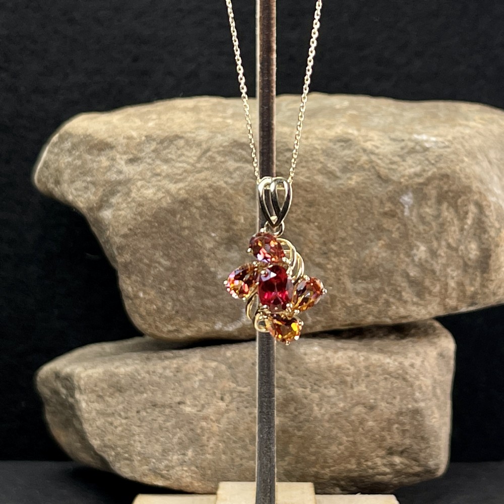 A 9ct yellow gold pendant featuring a central oval red garnet surrounded by four pear-cut orange garnets in a swirl setting, hanging from a gold chain. View II