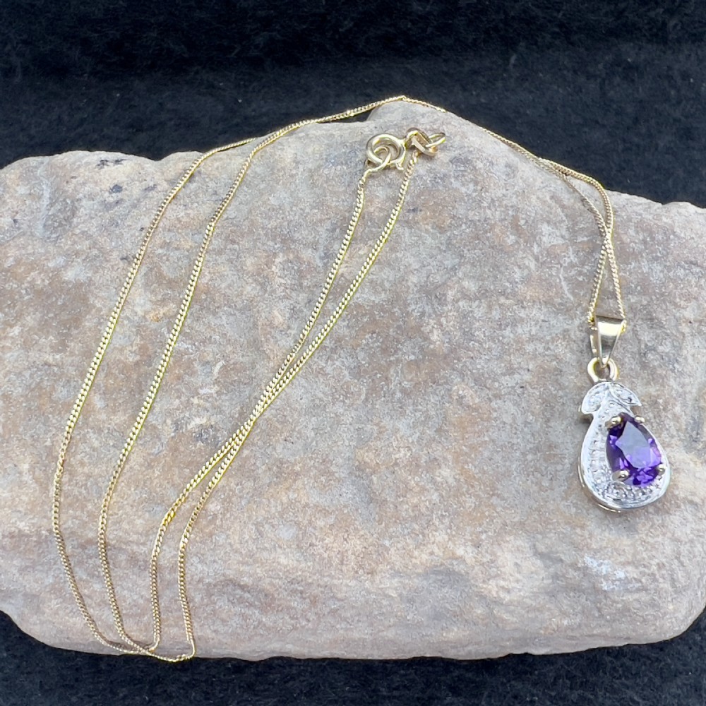 A 9ct gold teardrop pendant featuring a central oval purple amethyst set within a white metal border with accent diamonds, attached to a delicate gold chain. View XI