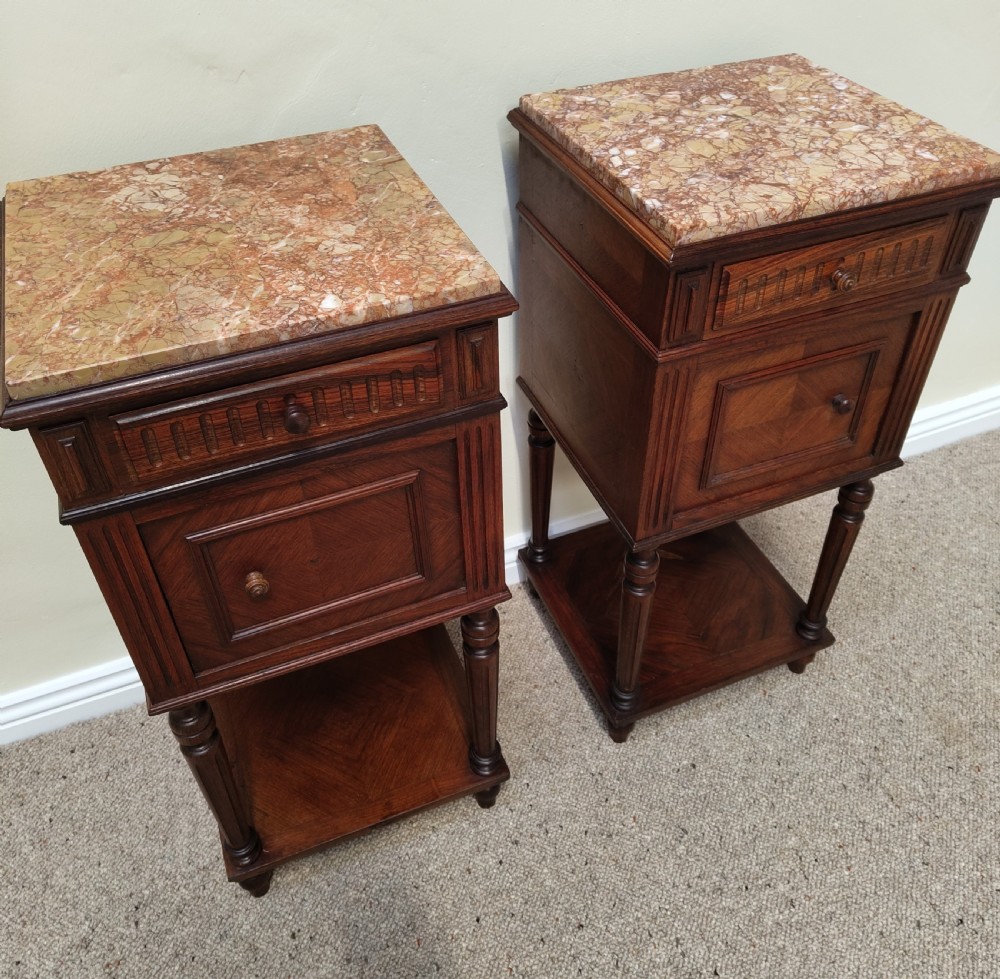 A pair of dark wood French bedside cabinets featuring carved drawer fronts, single cupboards, turned fluted legs, and variegated marble tops.
