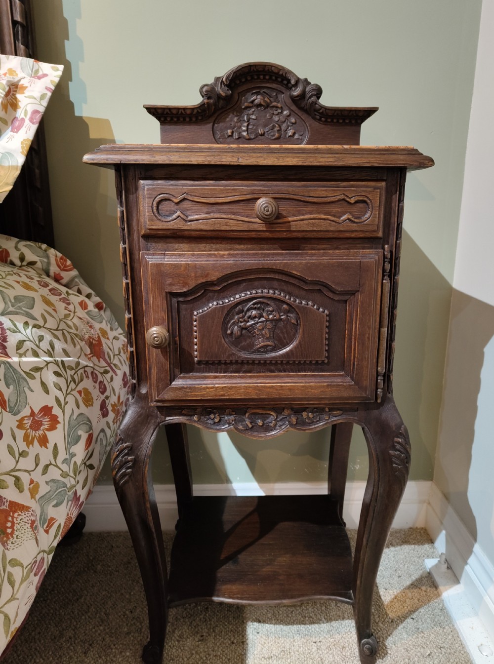 A dark oak French provincial bedside cabinet featuring a carved pediment top, a single drawer, a carved door cupboard, slender cabriole legs, and an open lower shelf.