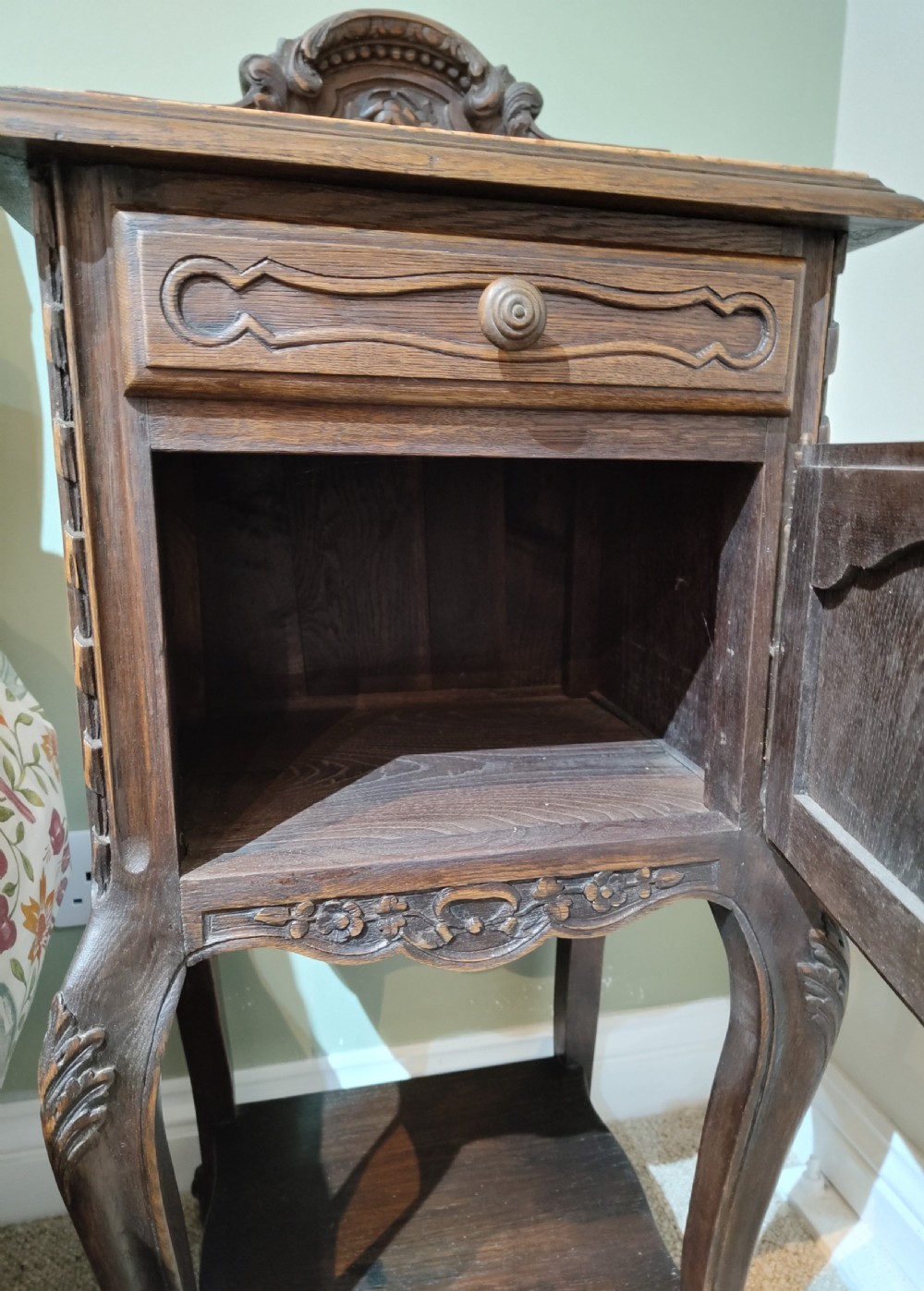 A dark oak French provincial bedside cabinet featuring a carved pediment top, a single drawer, a carved door cupboard, slender cabriole legs, and an open lower shelf. View IV
