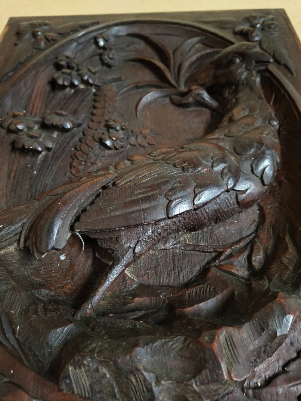 A rectangular dark wood panel featuring a high-relief carving of a bird surrounded by foliage within an oval frame. View II
