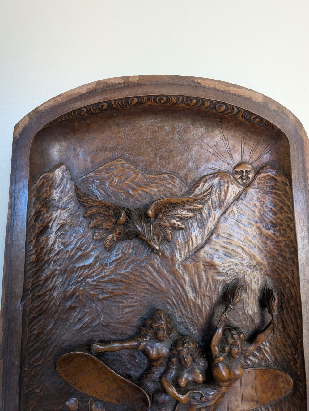 An arched carved wood wall panel featuring high-relief details of figures on a flying vessel, an eagle, a sun, and botanical elements in a warm brown finish. View II