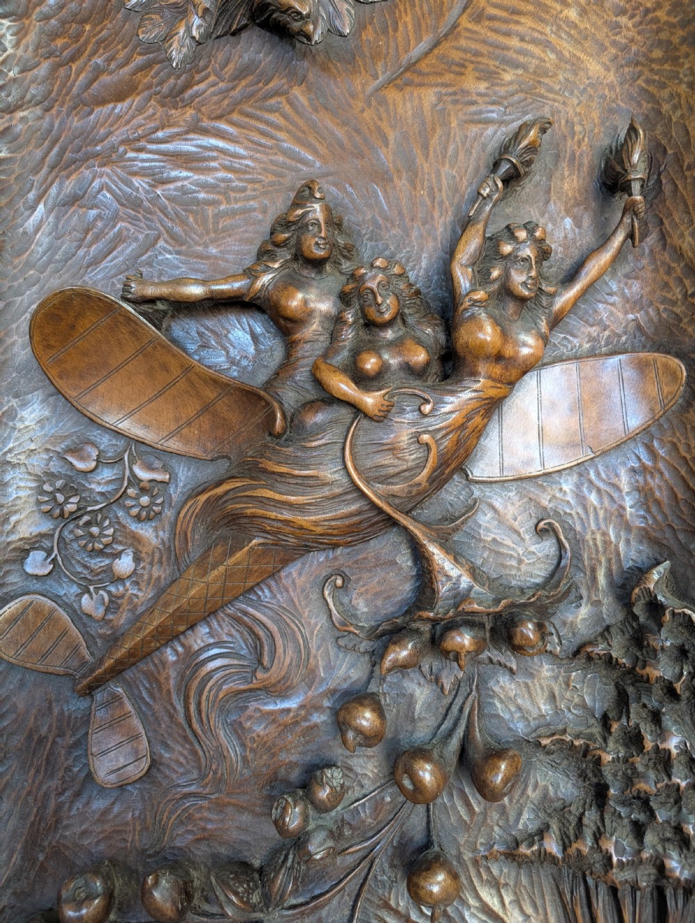 An arched carved wood wall panel featuring high-relief details of figures on a flying vessel, an eagle, a sun, and botanical elements in a warm brown finish. View III