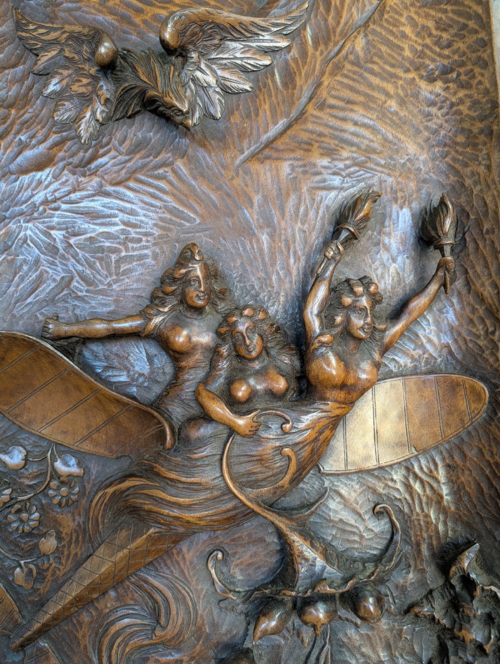 An arched carved wood wall panel featuring high-relief details of figures on a flying vessel, an eagle, a sun, and botanical elements in a warm brown finish. View VII