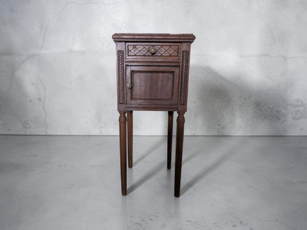 A tall dark brown wooden bedside cabinet featuring a carved drawer, a lower cupboard door, turned legs, and a marble top.