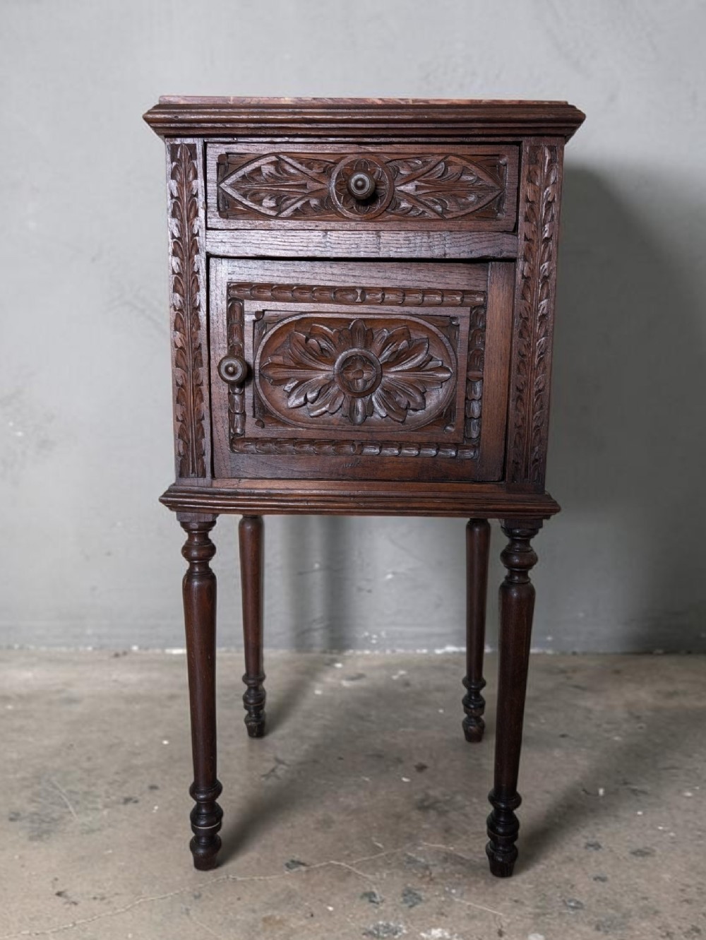 Carved dark oak bedside cabinet featuring a single drawer, cupboard door, marble insert top, and slender turned legs.