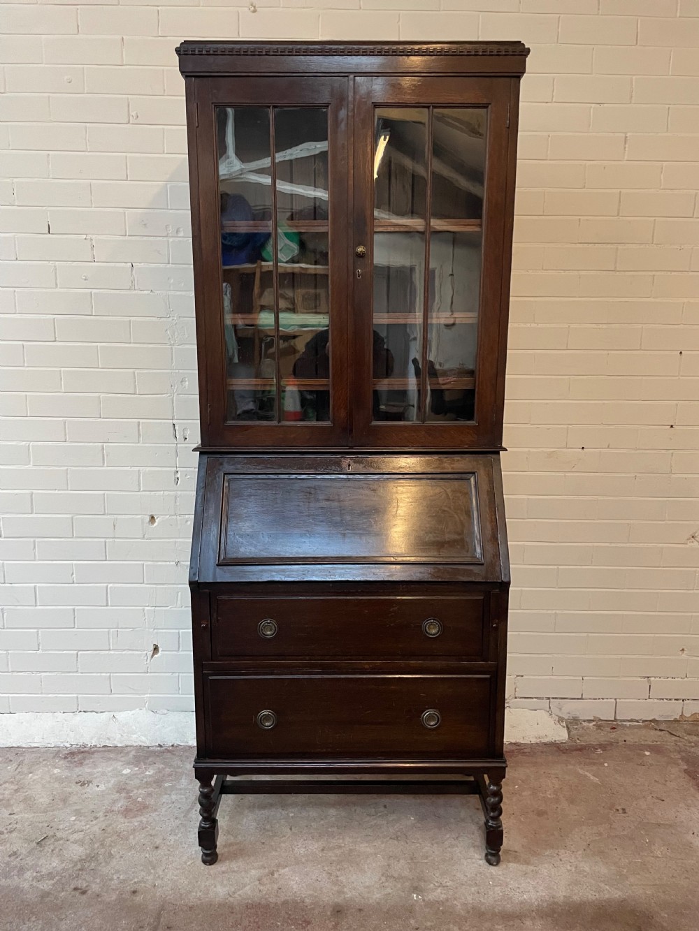 A dark oak Edwardian bureau bookcase featuring a twin-glazed door upper cabinet positioned above a slant-front writing slope and two lower drawers on turned legs.