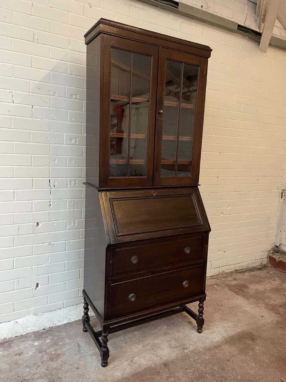 A dark oak Edwardian bureau bookcase featuring a twin-glazed door upper cabinet positioned above a slant-front writing slope and two lower drawers on turned legs. View II