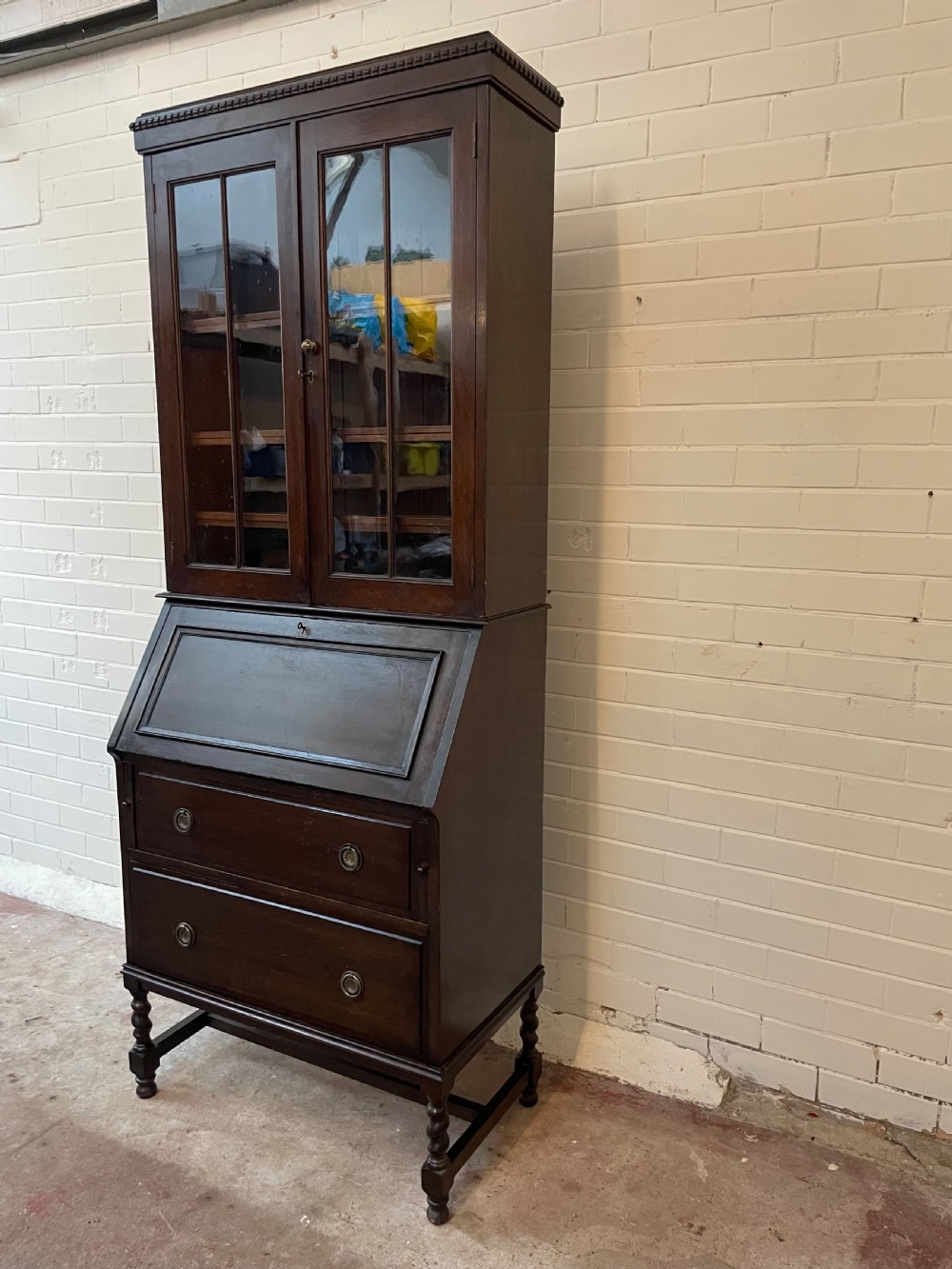A dark oak Edwardian bureau bookcase featuring a twin-glazed door upper cabinet positioned above a slant-front writing slope and two lower drawers on turned legs. View III